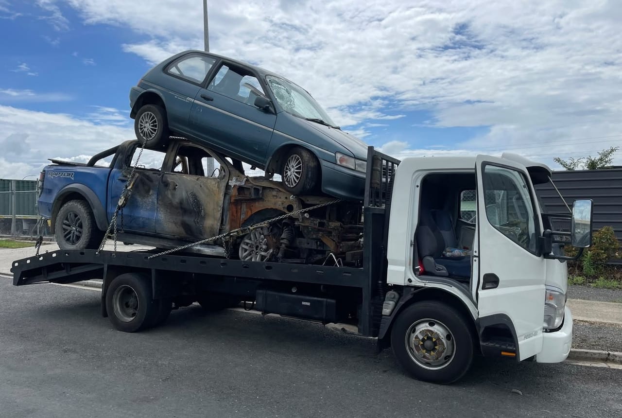 Flatbed truck loaded with two scrap cars including a burnt-out ute, removed in the Manawatū